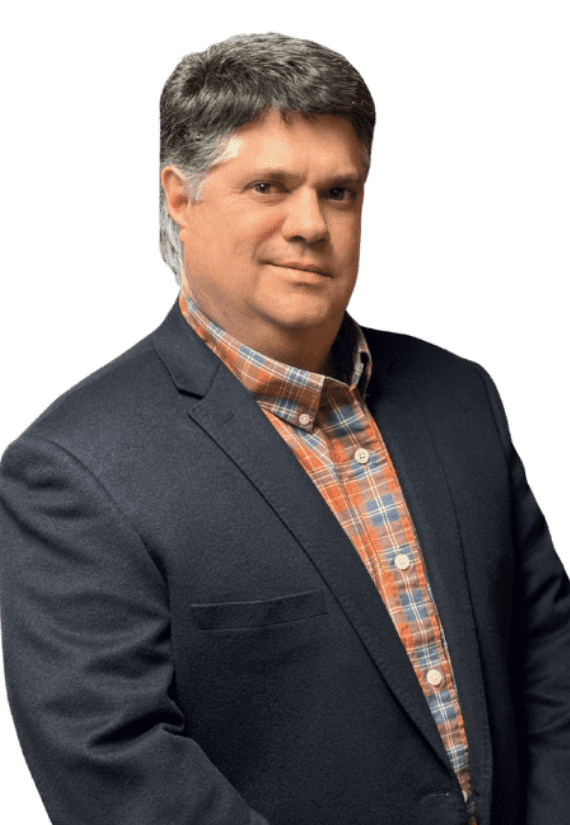 Man wearing a dark blazer over a plaid shirt, standing against a plain light background, looking at the camera with a neutral expression.